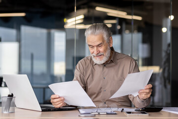 Senior businessman in a modern office reviewing paperwork at his desk with a laptop and reading glasses