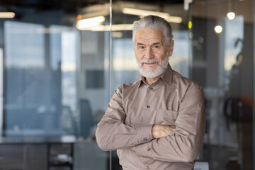 Confident senior businessman standing with arms crossed in a modern office setting, looking at camera.