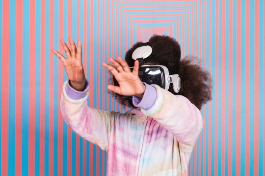 African American teenage girl in VR glasses experiencing virtual reality and touching air while standing on background of wall with rectangular tunnel