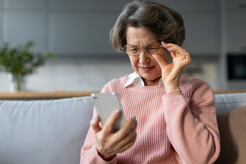 Confused senior woman in eyeglasses squinting eyes, having problems with vision, reading message on...