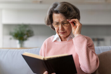 Senior woman squinting to see more clearly, touching eyeglasses, trying to read paper book, having...