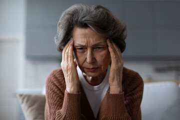 Stressed senior woman siting in living room, visibly distressed with severe headache, holding her temples in pain and discomfort