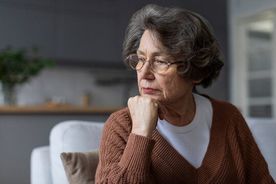 Pensive old woman sitting on couch at home alone, female deep in thoughts about health problems, suffer from sadness. Melancholy, loneliness, life on retirement