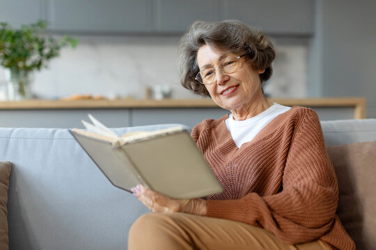 Aged woman sitting on couch and reading book pastime daydream, senior lady enjoying free time at home