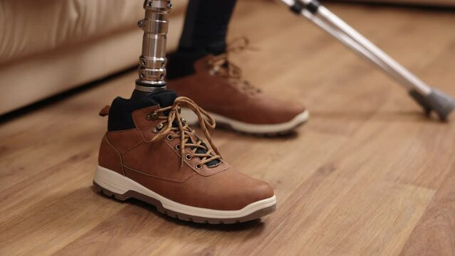 disabled man, rehabilitation program, inclusive technology. Close-up view of man with prosthetic leg sitting on sofa, putting on shoes with crutches beside him.