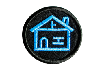 Round embroidered patch with blue house design on black fabric background, symbolizing home and community values.