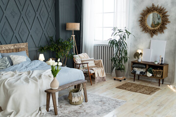 Stylish bedroom setup includes rustic wooden bed, dreamcatcher, and soft textiles. Wicker decor, indoor plants, and cozy armchair blend with serene ambiance. Bright window light enhances the space