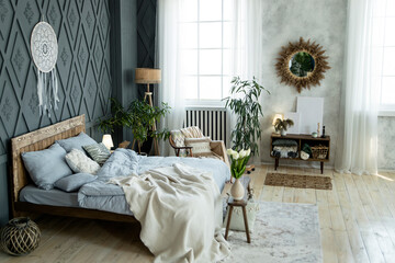 Modern bedroom with wooden bed, blue linens, wicker decor, and potted plants. Large window with white curtains provides natural light. Dreamcatcher and rustic elements enhance the cozy atmosphere