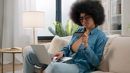African American ethnic girl making online payment holding bank credit card using modern laptop at...
