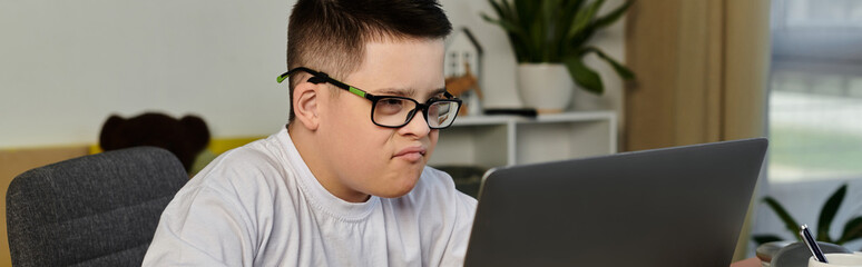 A young boy with Down syndrome sits at a desk, intently looking at his laptop.