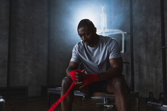 Fitness workout in gym. African American man fighter preparing for fight wrapping hands with red boxing wraps sports protective bandages in gym. Strong man ready for fight boxing sparring training