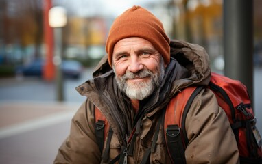 Fototapeta premium A man wearing a brown jacket and orange hat is smiling. He is sitting on the sidewalk with a backpack on his back