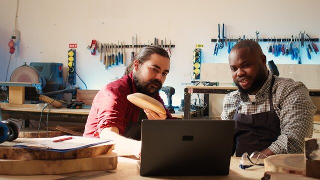 Carpenter and colleague verifying finished wood art product, comparing with blueprint. Artisan and apprentice inspecting wooden object for damages based on schematics on laptop screen, camera B
