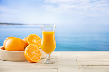 Glass of orange juice and oranges cut in halves on wooden table top with sea background. Drinks on summer hot day.