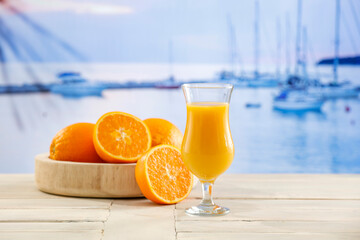 Glass of orange juice and oranges cut in halves on wooden table top with sea background. Drinks on summer hot day.