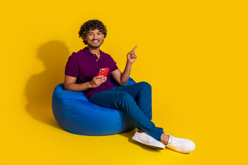 Full body photo of young man sit bean bag hold device point empty space dressed stylish violet...