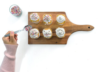 Closeup woman hand holding spoon with multicolor frosting, decorating cupcake on a wooden board, adding a colorful touch to the cupcakes' presentation. Top view