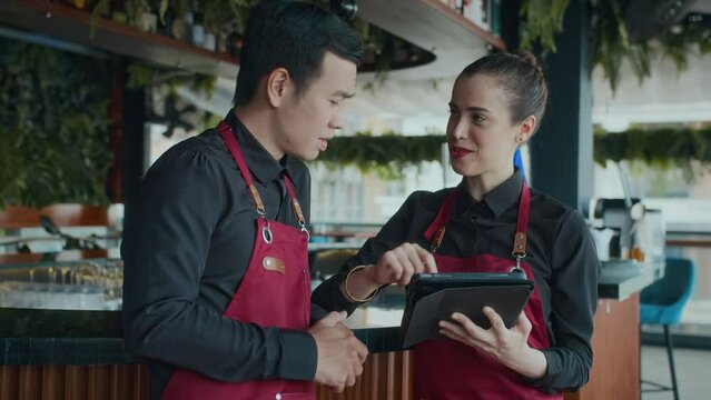 Medium shot of Caucasian waitress instructing Asian male newcomer using tablet computer at restaurant