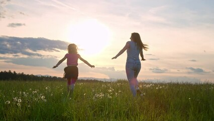 Slow motion shot of a joyful mother and her little daughter in the green meadow running with open arms at sunset, rear view. Life happiness concept.