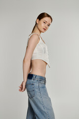 A young woman poses against a white backdrop, wearing a white top and denim jeans.