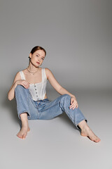 A young woman poses confidently in a white corset top and blue jeans against a gray backdrop.