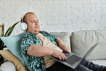 A man with inclusivity wearing headphones sits on a couch, working on a laptop.