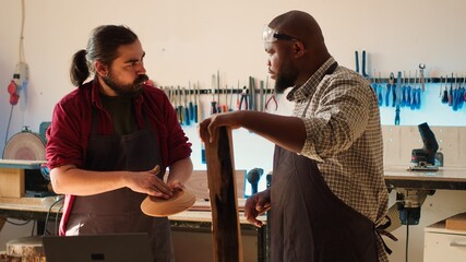 Woodworker looking over blueprints on laptop, brainstorming with colleague next steps in wood processing. Manufacturer and BIPOC apprentice reading furniture assembly schematics in studio, camera A