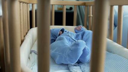 Newborn baby in a blue outfit resting in a wooden crib, viewed through the crib bars, symbolizing the safety and protection provided to newborns in a hospital setting