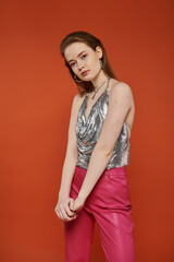 Fototapeta premium Young woman in chainmail top and pink pants poses against orange background.