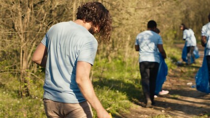 Team of volunteers cleaning the forest area from garbage and plastic, collecting rubbish with tongs and bags. Activists doing voluntary work to preserve natural habitat, illegal dumping. Camera A.