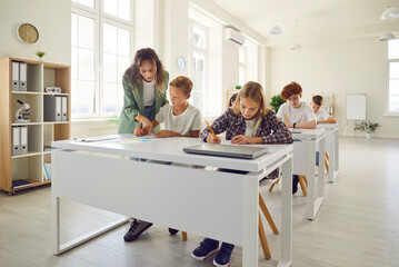 Group of children sitting at desks in the classroom and writing in their notebooks. Woman school...
