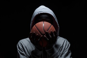 Mysterious figure in hoodie holding basketball, shadows and light playing across face emphasizing the sport's intensity and style.