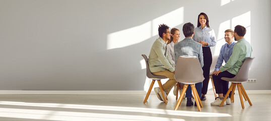 Business coach meeting with a team of young people. Young woman talking to a group of happy diverse men and women sitting in a circle in a modern office with a grey copy space wall. Banner background