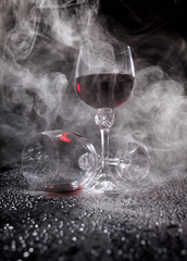 Two glasses on a black background. Smoke around a glass of wine. A glass of red wine stands on the surface, an empty glass lies next to it.
