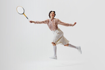 Elegant young beautiful woman in vintage style clothes and hairstyle in motion with tennis racket, playing isolated on white background. Concept of sport, retro and vintage, active lifestyle, hobby