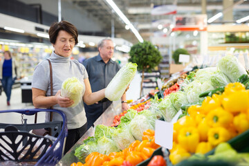 Elderly woman examines china cabbage in vegetable section to choose juiciest cabbage
