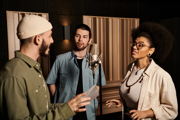 Three individuals converse animatedly in a recording studio, preparing for a music band rehearsal.
