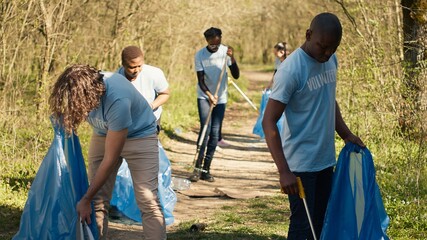 Diverse men volunteers pick up rubbish and plastic trash with tongs, working to combat illegal dumping and preserve natural forest environment. Activists volunteering for litter cleanup. Camera A.