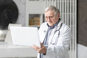 Senior doctor sits at the desktop and works on laptop in his modern medical office. Online consultation concept