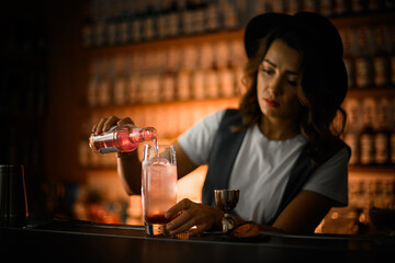 Female bartender is preparing a cocktail, pouring a pink drink from a bottle into a glass with ice