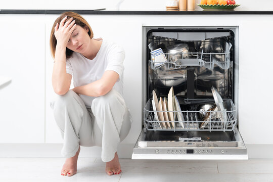 Tired and bored woman putting dirty plates in dishwasher machine in the kitchen. Household and exhausting cleaning day concept
