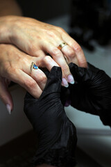 A manicurist wearing black gloves gently holds a client’s hand while performing nail care, showcasing professional and meticulous attention to detail in a salon setting.