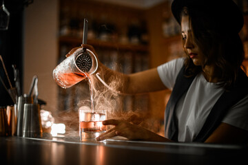 Woman bartender in a white t-shirt and vest pours a cocktail with ice from a mixing glass into another glass