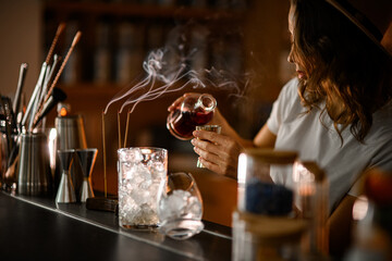 Bartender pours a brown drink from a bottle into a jigger, glasses and smoking incense sticks stand on the bar counter