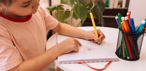 Kid draws with a felt-tip pen with her left hand in a notepad on a white table.