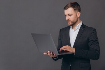 Portrait of successful and confident businessman in grey suit, working with laptop, standing over...