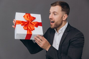 Bearded happy smiling man in business suit holding a gift and unwrapping it on a gray background,...