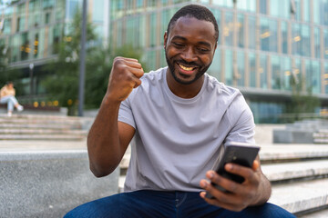 Happy African American man euphoric winner with smartphone on street in city. Person guy looking at...