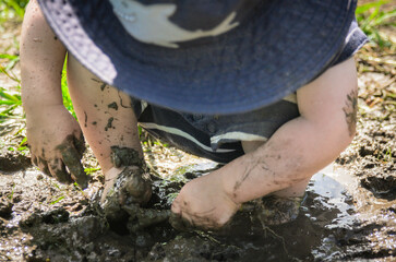 Toodler playing in the mud with their hands, wearing a blue hat.
