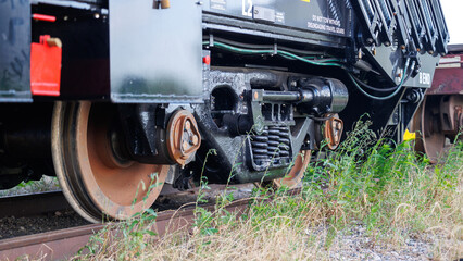 The close-up of the detailed mechanics of a freight train's wheel and suspension system. Train sits on tracks with a grass and weeds in front.